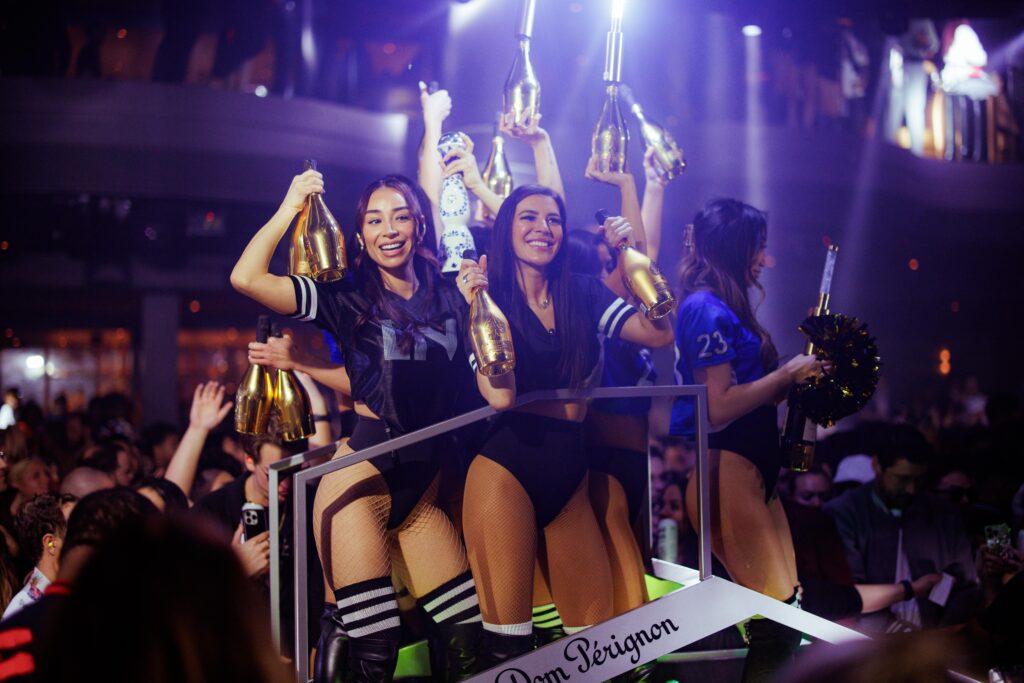 Group of bottle servers standing on top of a vessel in the GA crowd holding Dom Perignon Rose Gold champagne bottles and Clase Azul Tequila Reposado bottles with lights. They are wearing LIV football style jerseys smiling with one server holding a pom pom at the front of the vessel.