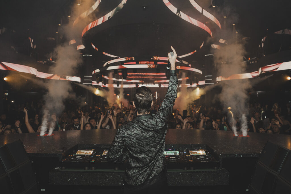Angle from behind the DJ booth of Sam Feldt's back pointing up to sky looking into the GA crowd
