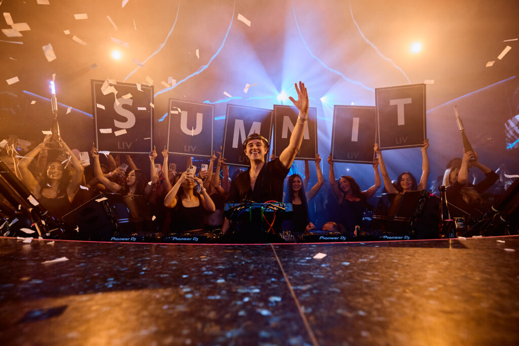 Front facing angle of John Summit smiling with his arm in the air looking toward the crowd and bottle servers holding letter boards that spell out "SUMMIT" behind him