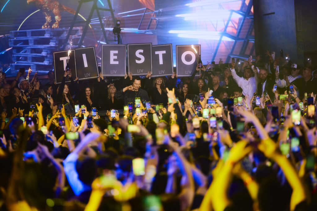 Taken from the crowd, Tiësto holding his arm up with bottle servers holding letter boards that spell out "TIESTO" behind him