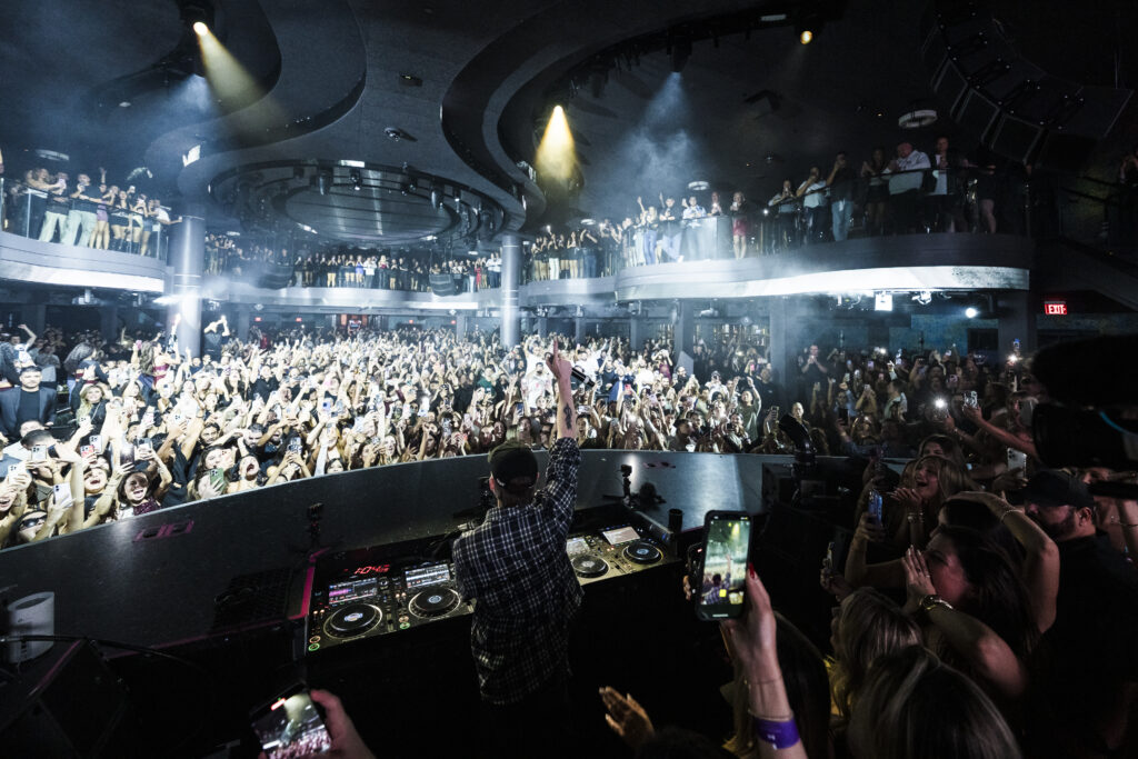 Angle taken from behind Dom Dolla in DJ booth with his arm up facing the crowd