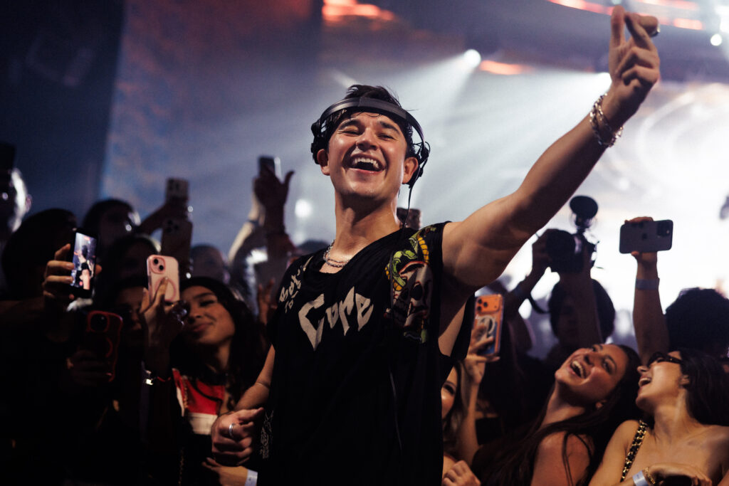 Closeup image of John Summit behind the DJ booth, with one arm raised, smiling at the crowd. 