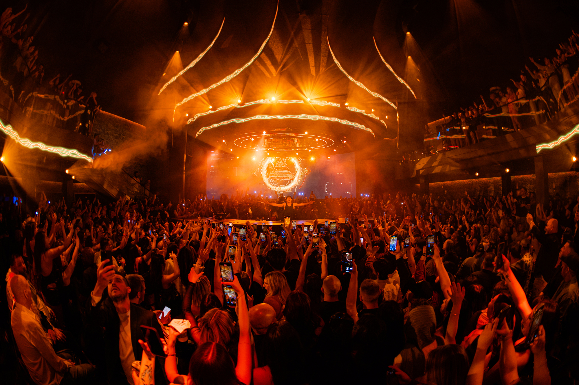 John Summit behind the DJ booth with his arms open, performing to a fully packed venue.