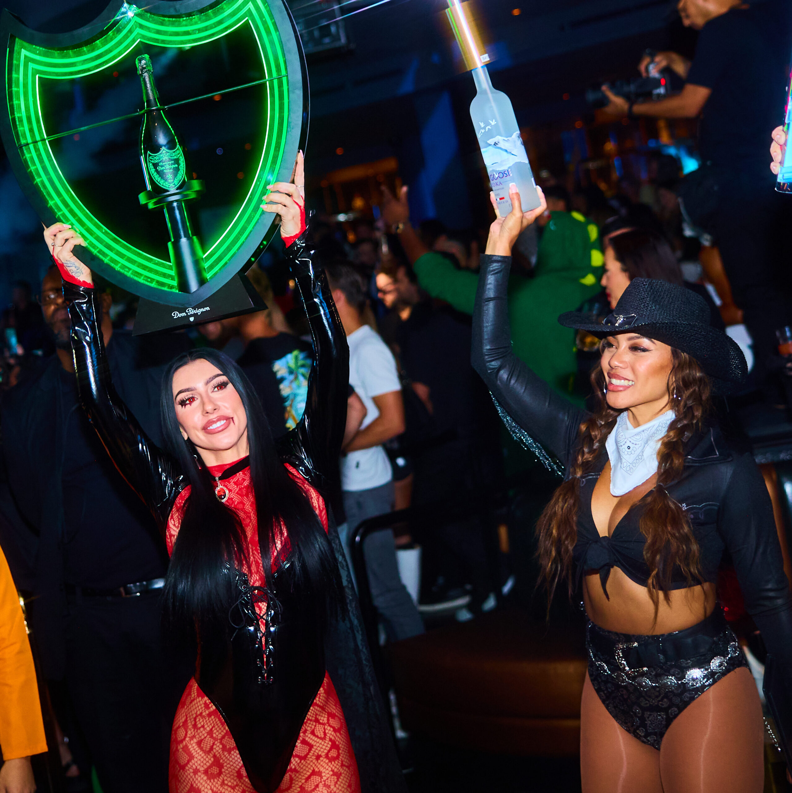 Two servers holding bottle presentations of vodka and champagne, dressed in Halloween costumes and dancing.
