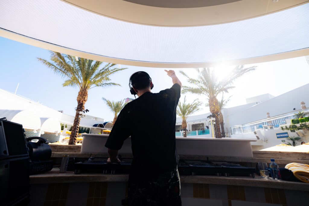 Cloonee behind the DJ booth with one arm raised and palm trees in the background. 