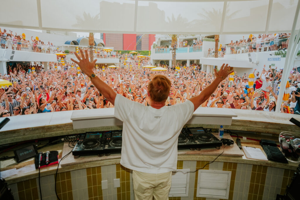 David Guetta facing the full crowd behind the DJ booth and raising his arms.