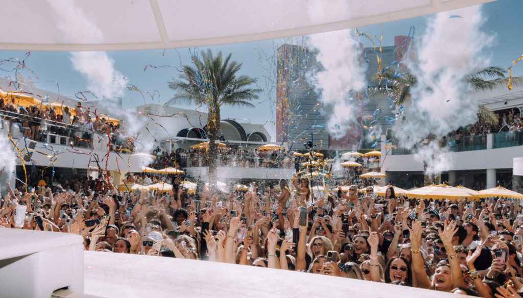 Crowd celebrating at LIV Beach with confetti cannons, palm trees, and packed daytime pool party atmosphere.