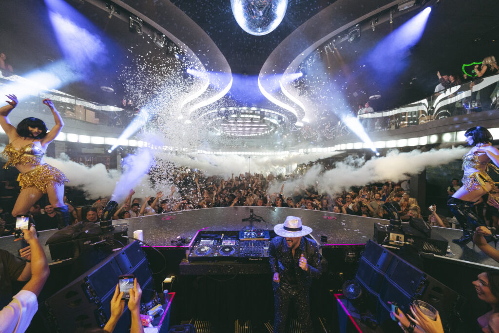 Disco Dom dancing behind the DJ Booth at LIV Las Vegas with confetti, disco ball, and packed crowd.