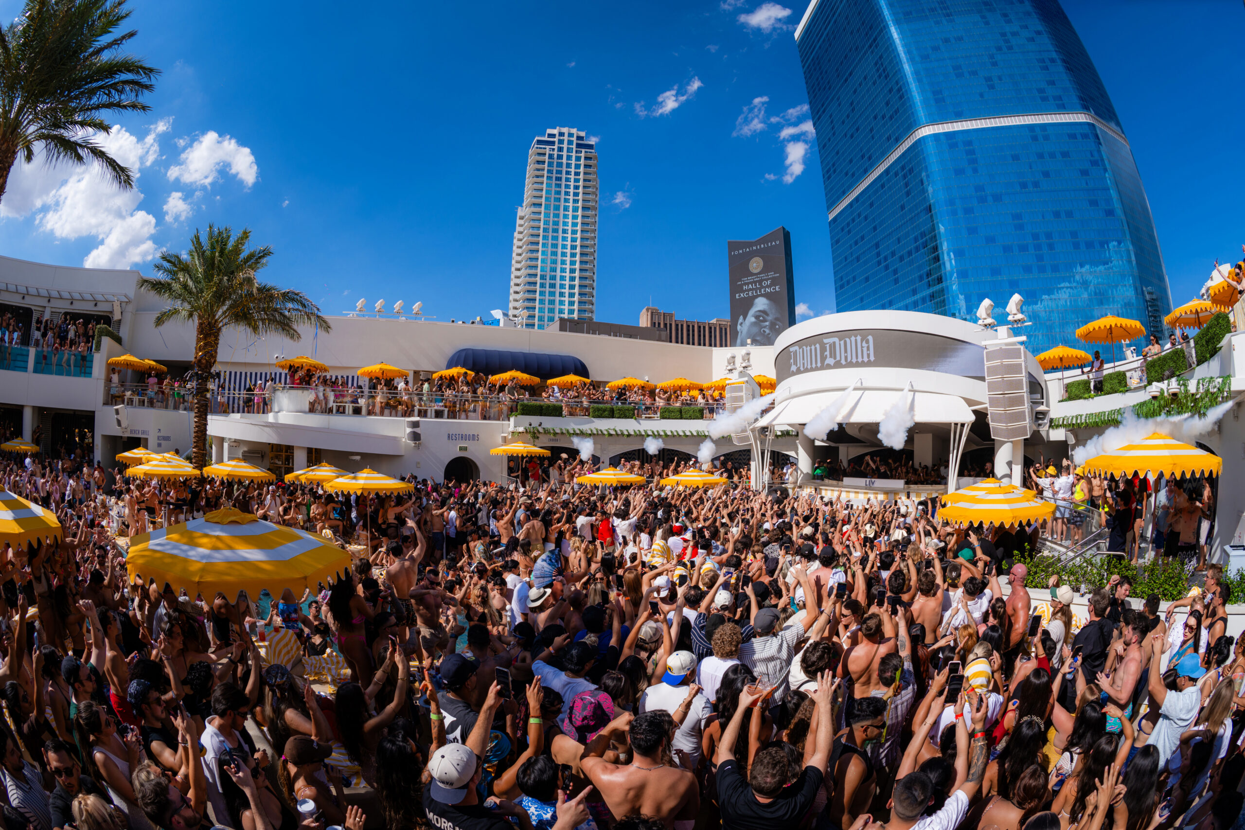 Crowd packed around the LIV Beach DJ booth as smoke cannons fire during a high-energy daytime set, with "Dom Dolla" displayed on the LED.