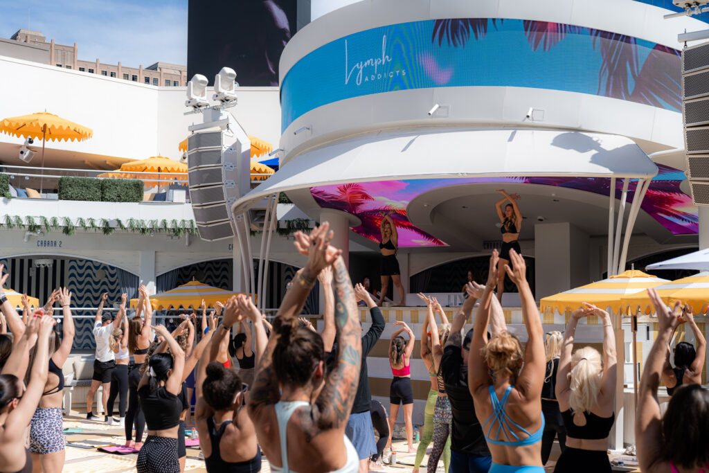 Large group fitness session at LIV Beach with instructor-led movement in front of the DJ booth and LED screens displaying "Lymph Addicts".