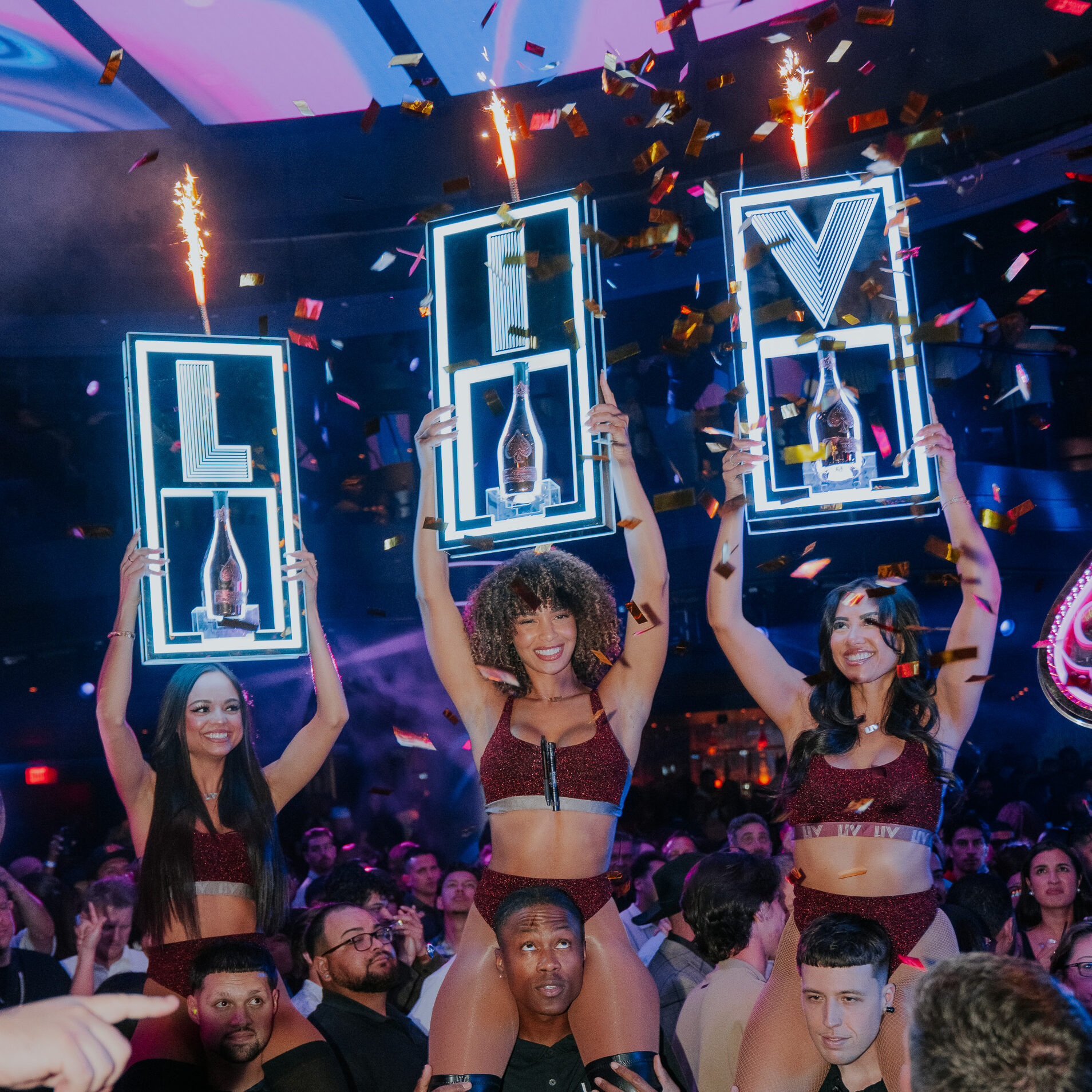 Model cocktail servers holding champagne bottles with LED signage that spells "LIV".