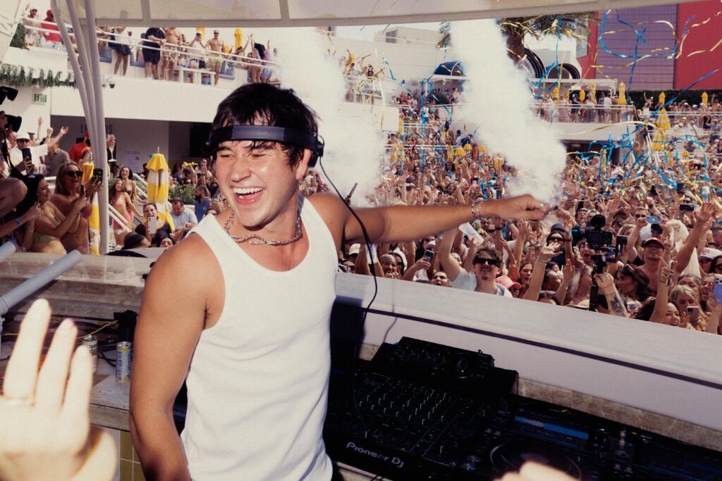John Summit dancing with one arm raised behind the DJ booth, with confetti.