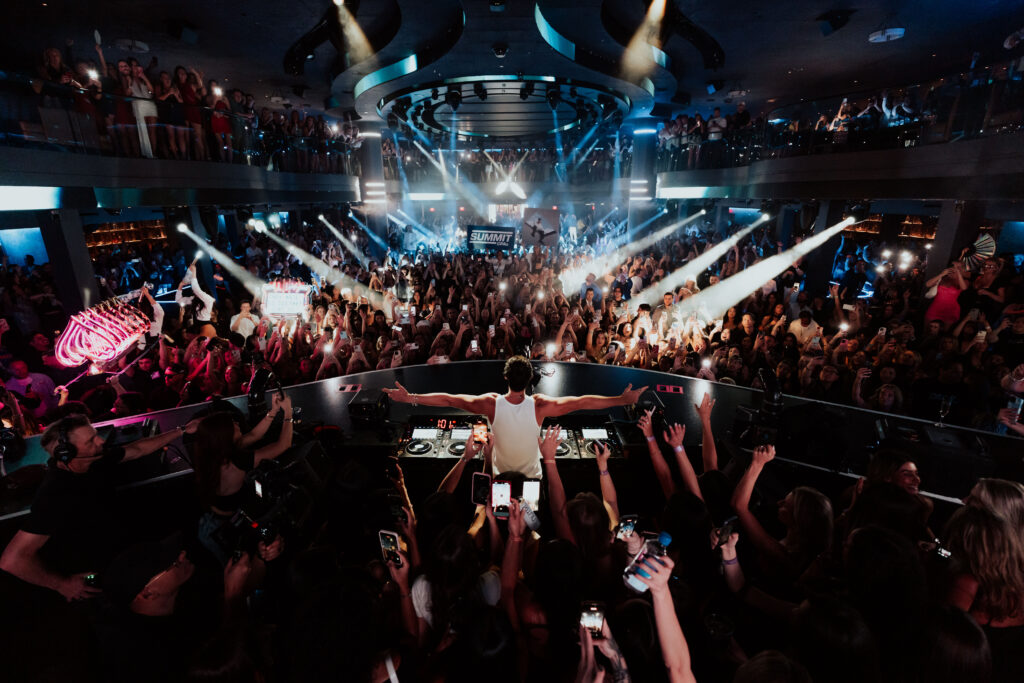 John Summit behind the LIV Nightclub DJ booth with his arms open to the packed audience.