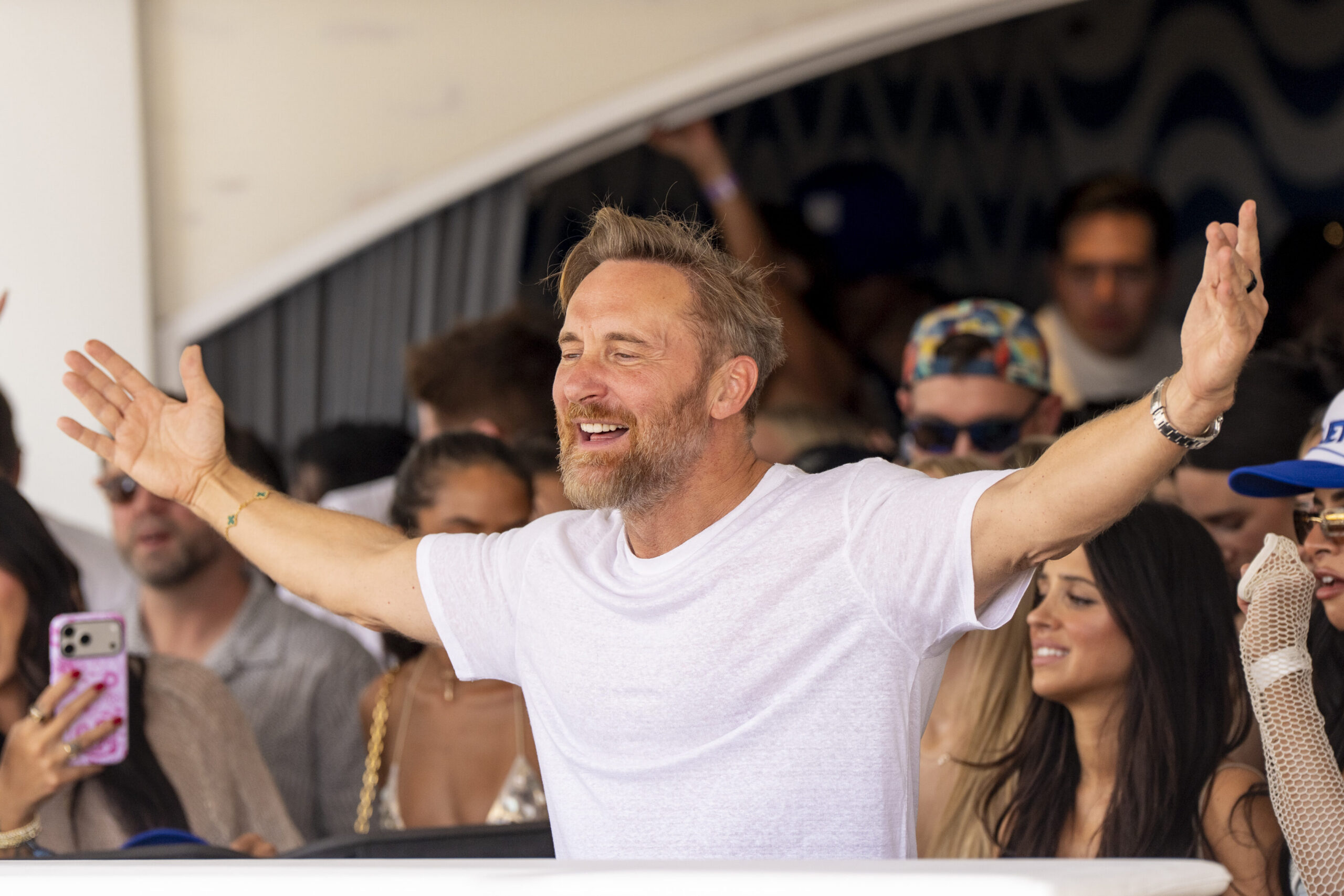 David Guetta with his arms raised, facing the crowd from the DJ booth.